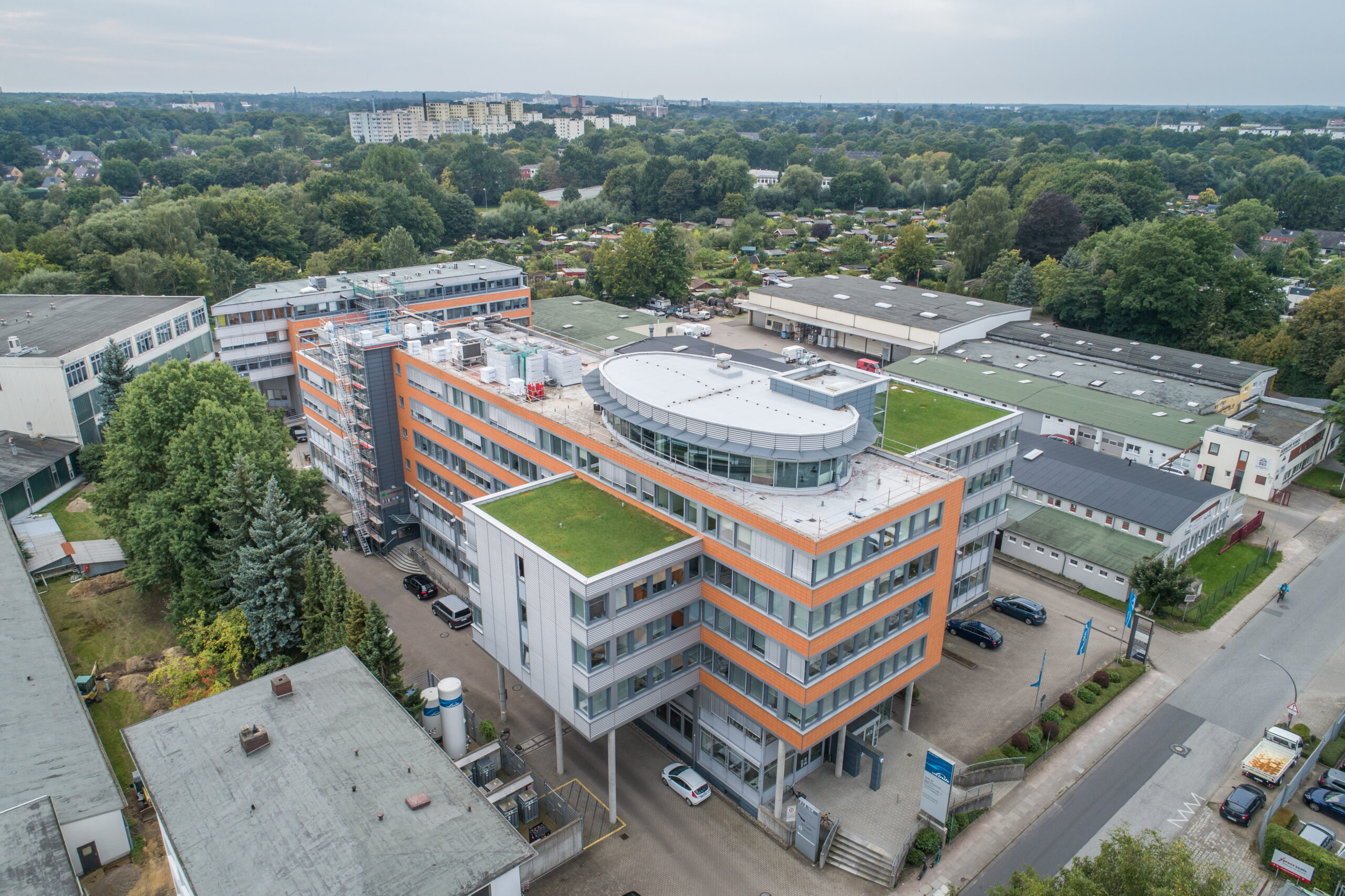 Aerial view of a modern multi-story building with a green roof, surrounded by trees and other buildings, set in an urban environment.