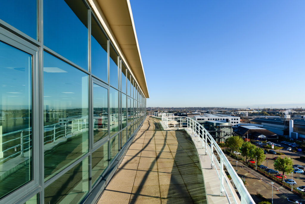 View of a modern building's glass facade reflecting a clear blue sky, with a paved balcony and distant cityscape.