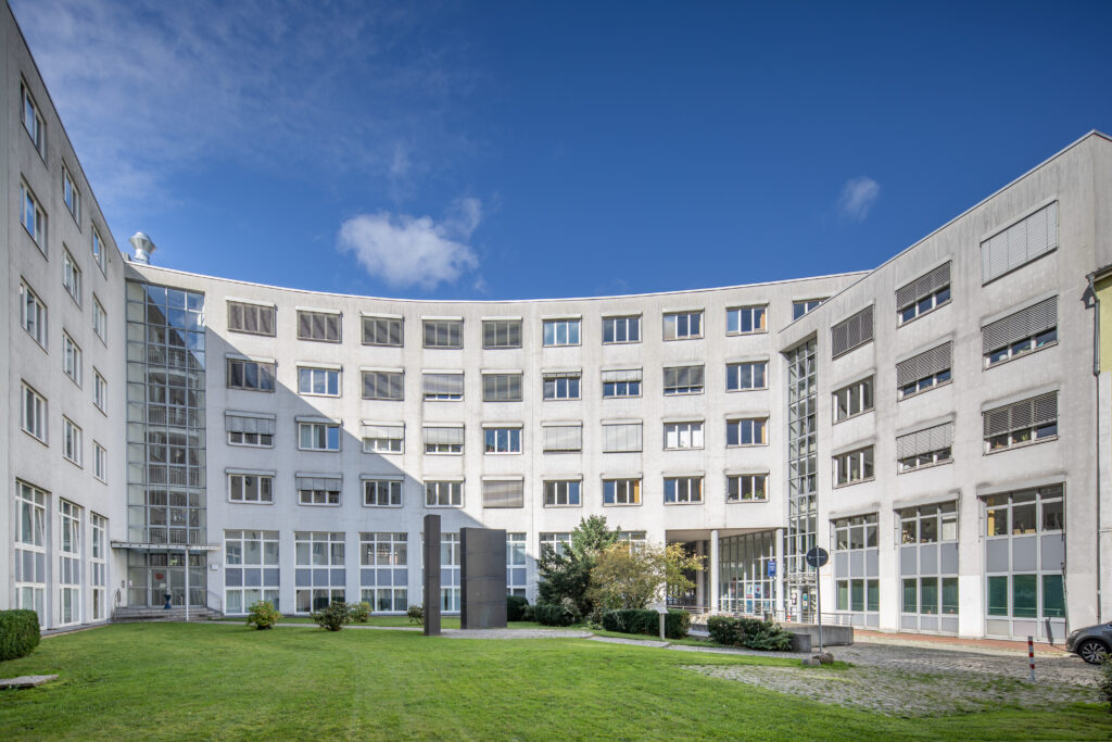 Modern office building with a curved design, featuring large windows and a grassy courtyard in the center, under a clear blue sky.