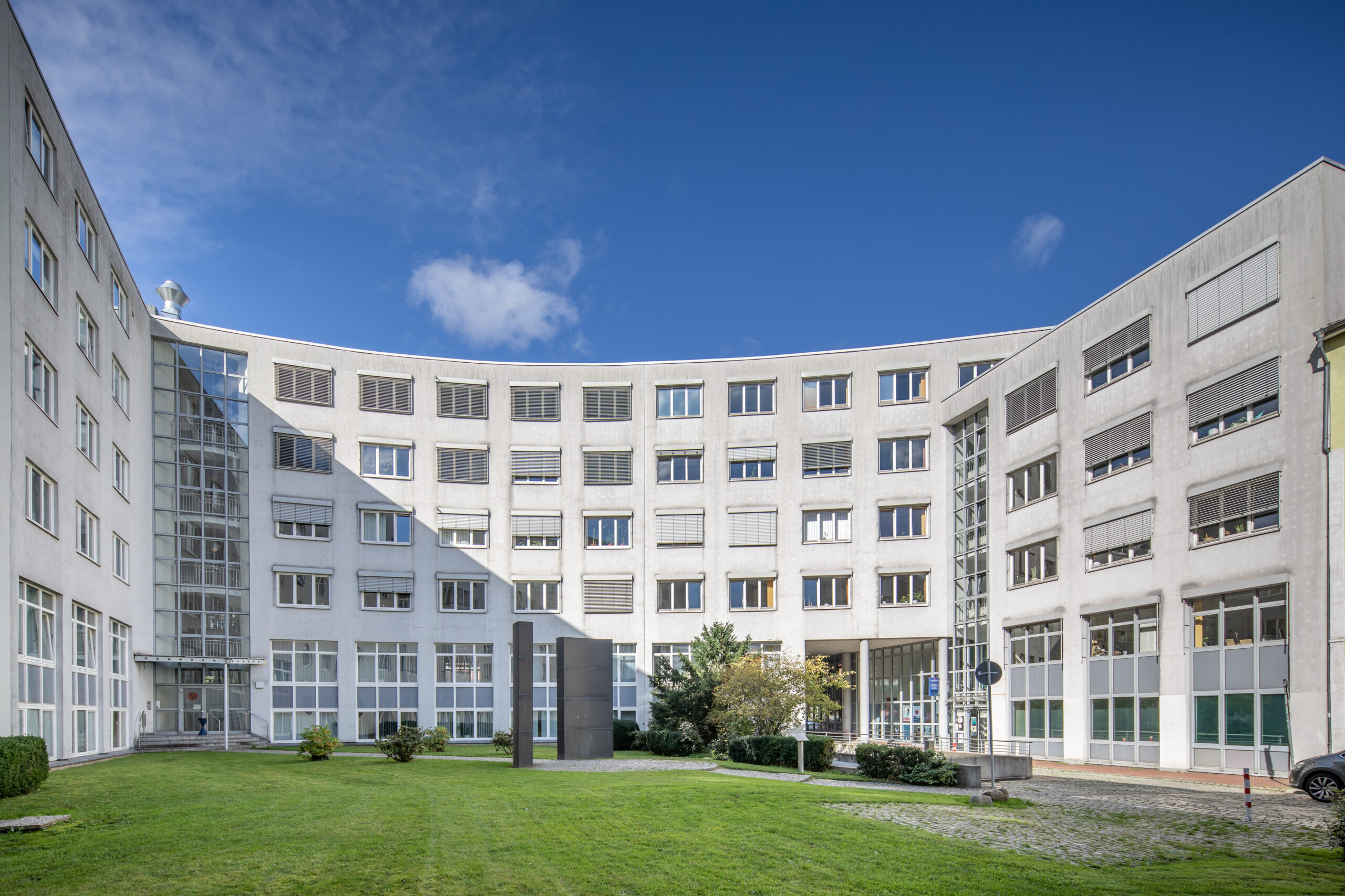 Modern office building with a curved design, featuring large windows and a grassy courtyard in the center, under a clear blue sky.