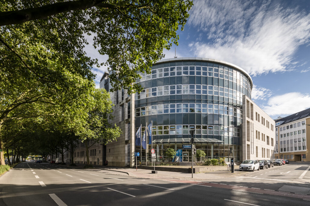 Modern circular office building with glass facades, surrounded by trees and empty streets, under a partly cloudy sky.