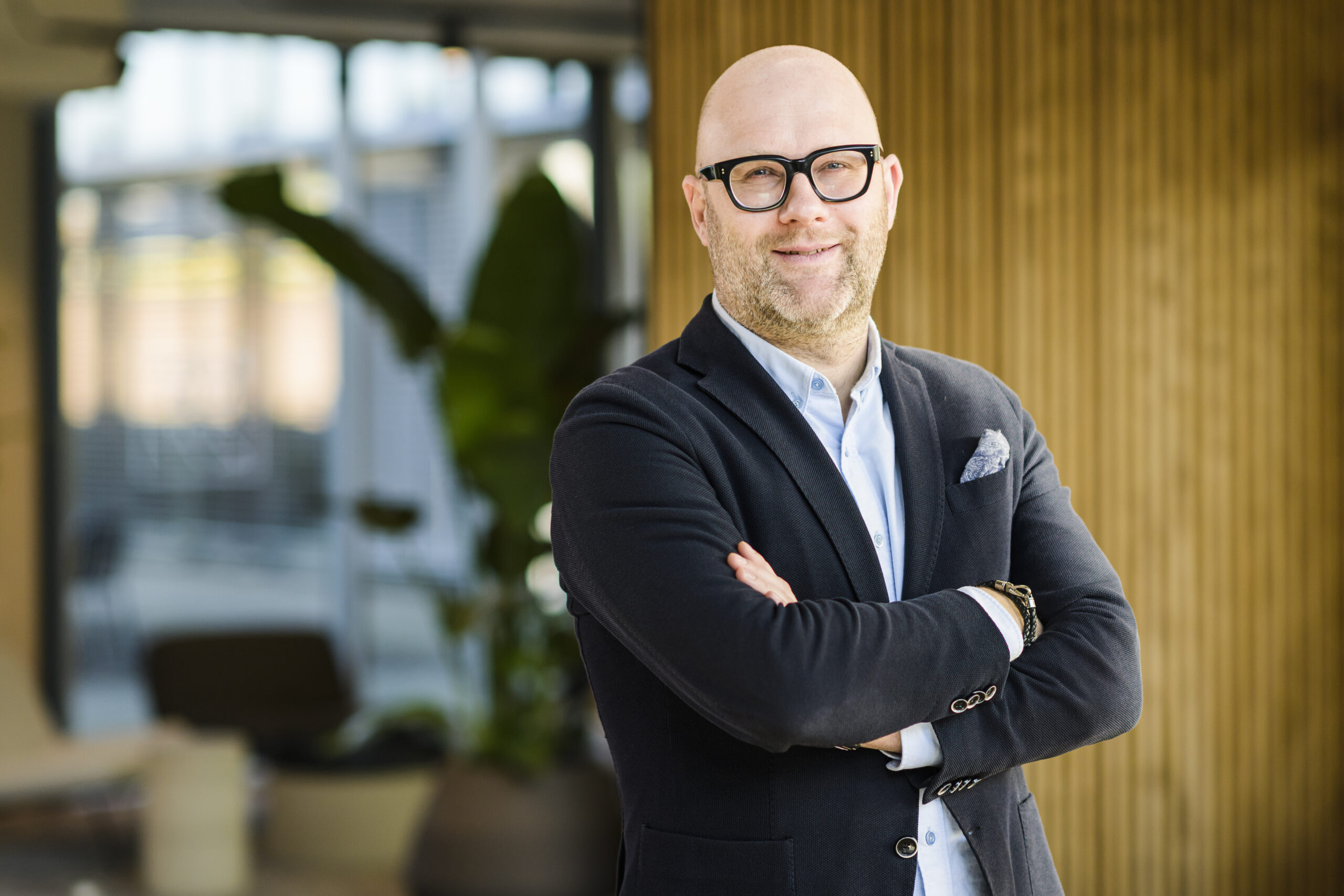 A bald man with glasses stands confidently in an office setting, wearing a black blazer and light blue shirt, with arms crossed.