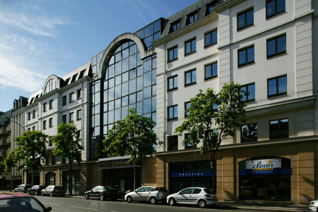 Street view of modern office buildings with large windows. Several trees line the sidewalk, and cars are parked along the street. Various businesses are on the ground floor.