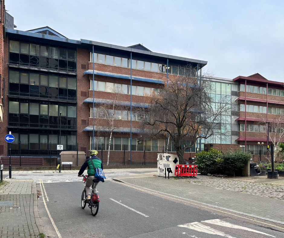 Person on a bicycle rides down a street lined with multi-story brick and glass buildings under a cloudy sky.